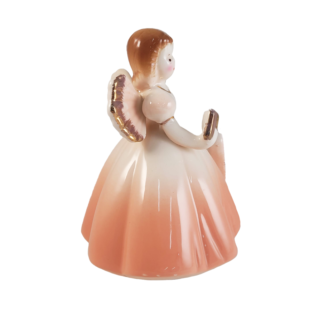 Side of porcelain angel figurine with a peach gradient dress holding a book on a white background