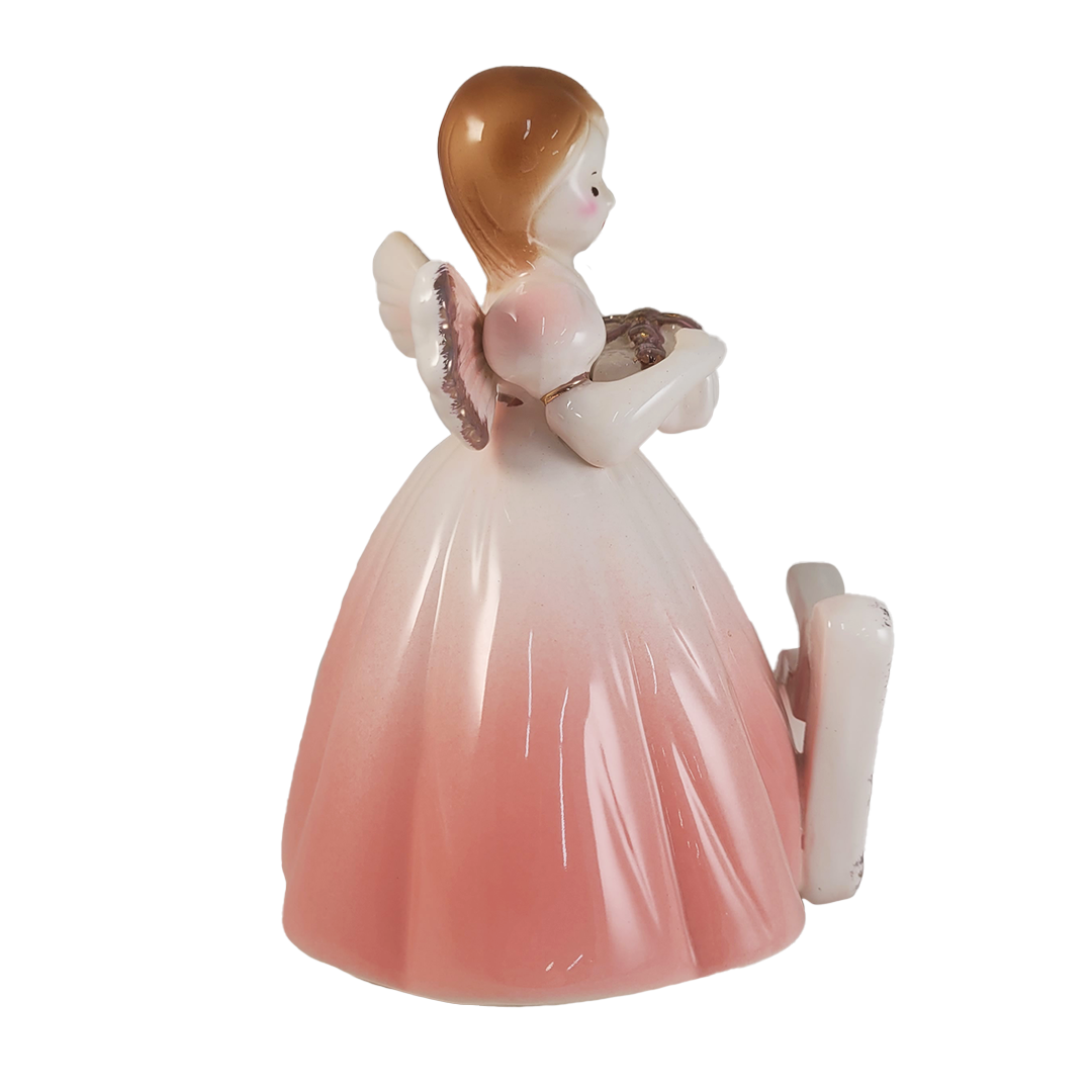 Side of angel figurine holding a gift wearing a light pink dress on a white background