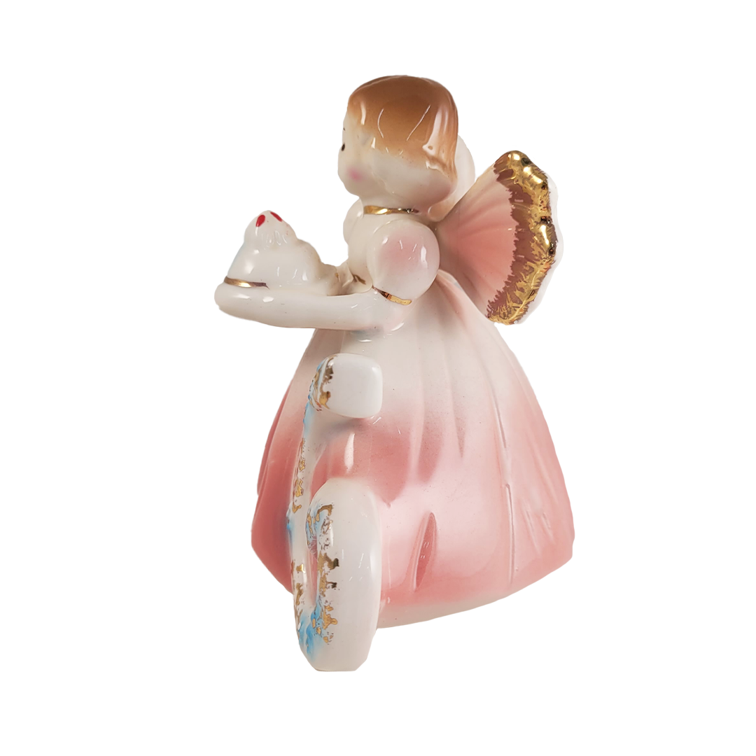 Side of porcelain angel figurine with a pink dress and gold wings holding a cake on a white background