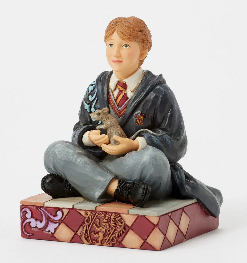 Quarter view of figurine of Ron Weasley in a robe holding a rat, sitting on a box with Gryffindor emblem.