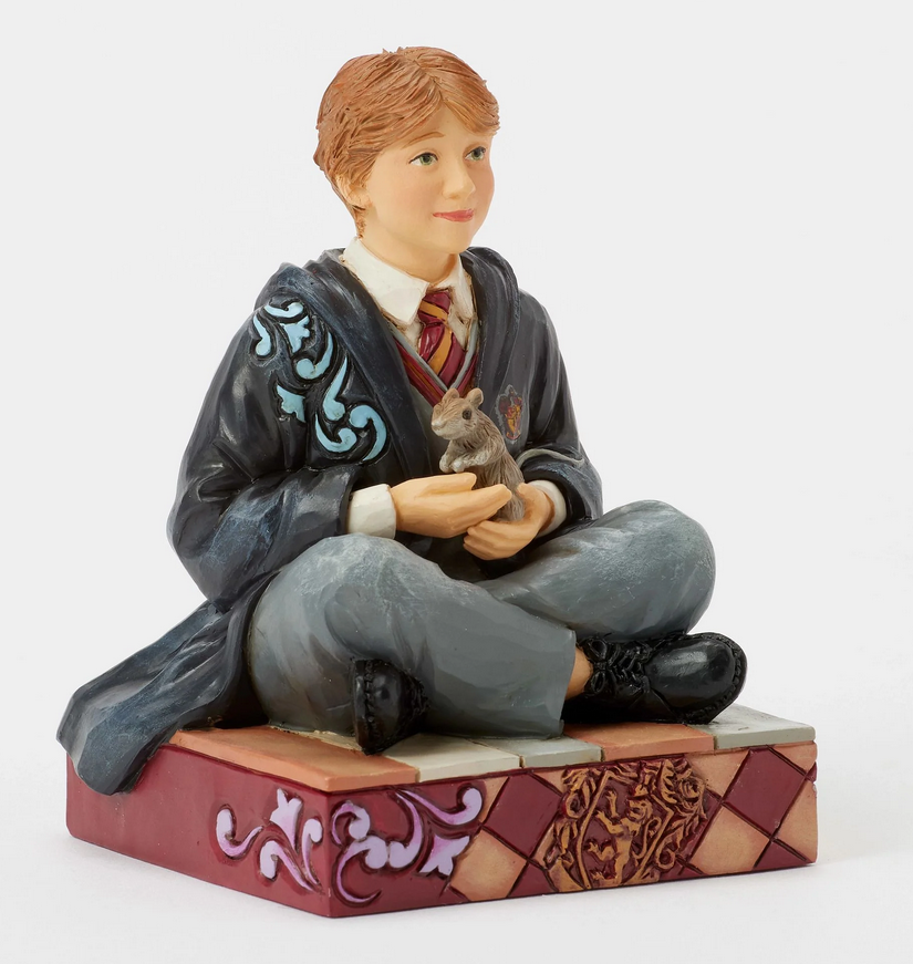 Quarter view of figurine of Ron Weasley in a robe holding a rat, sitting on a box with Gryffindor emblem.