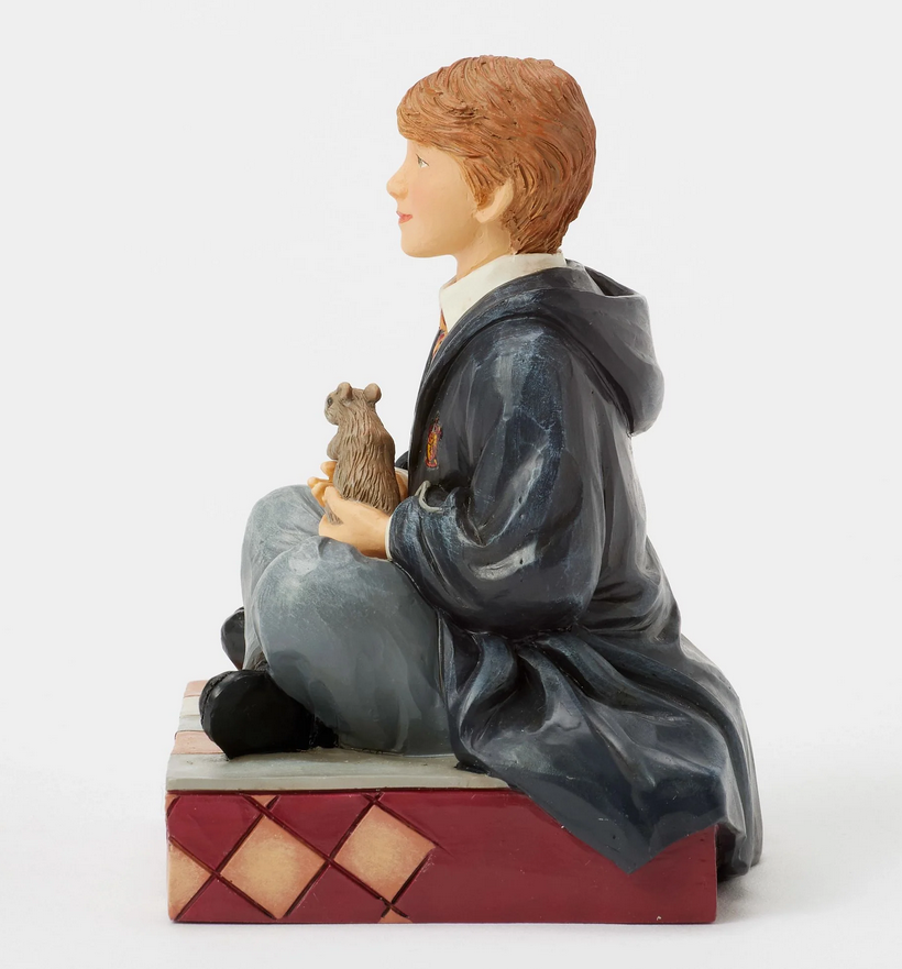 Left side of figurine of Ron Weasley in a robe holding a rat, sitting on a box with Gryffindor emblem.