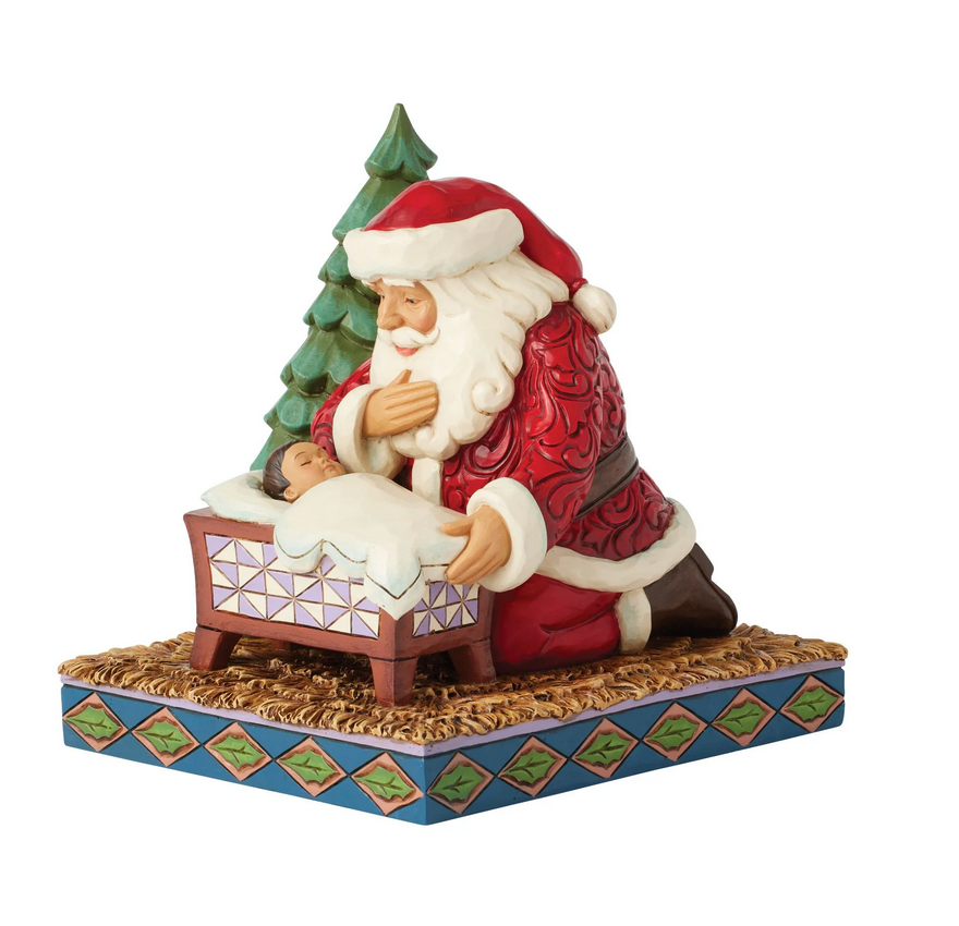 Santa Claus figurine looking at a baby in a manger with a Christmas tree on a white background