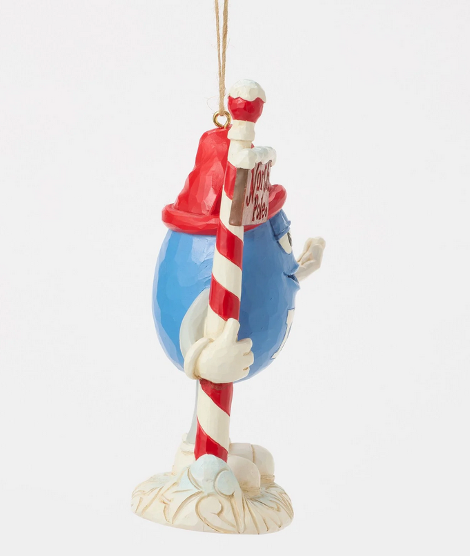Side of decorative ornament of a blue M&M character with a red hat holding a candy cane north pole sign on a white background