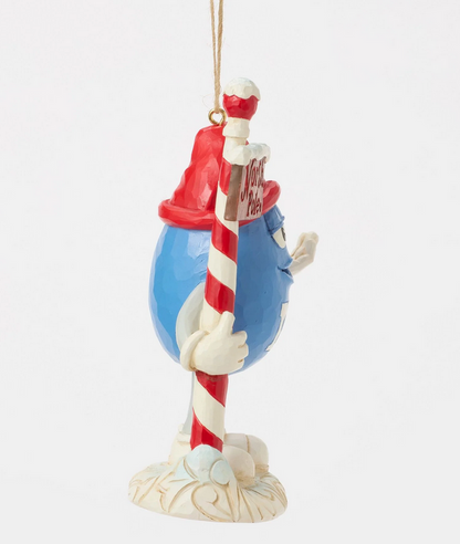Side of decorative ornament of a blue M&M character with a red hat holding a candy cane north pole sign on a white background