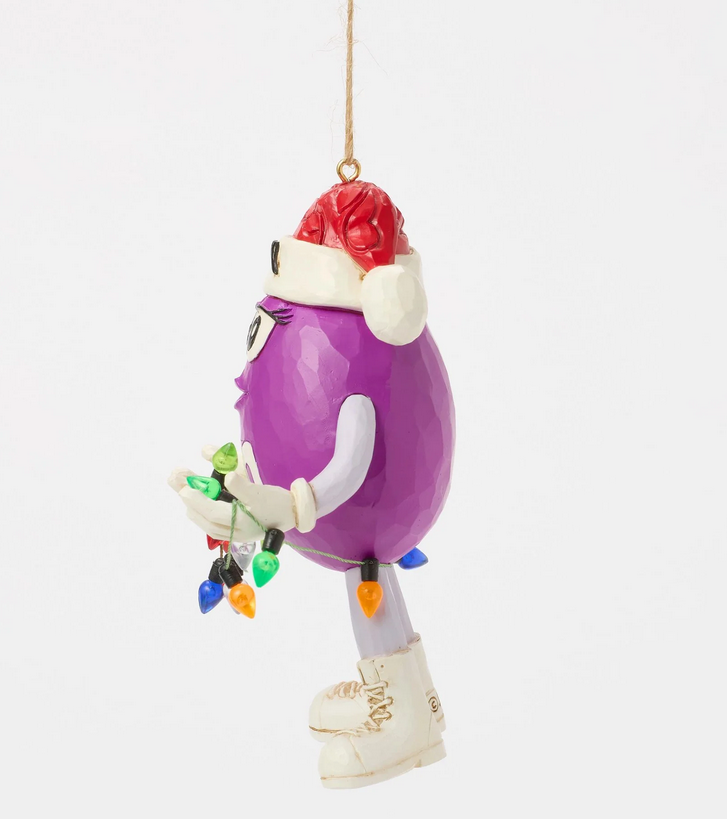 Left side of purple M&M character ornament wearing a Santa hat holding Christmas lights on a white background