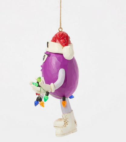 Left side of purple M&M character ornament wearing a Santa hat holding Christmas lights on a white background