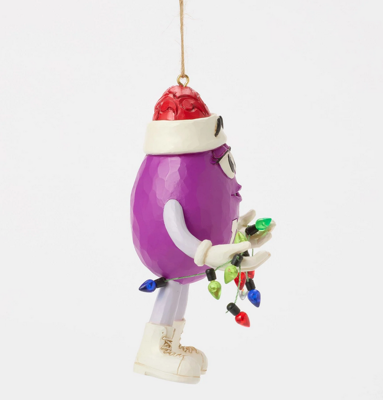 Side of purple M&M character ornament wearing a Santa hat holding Christmas lights on a white background