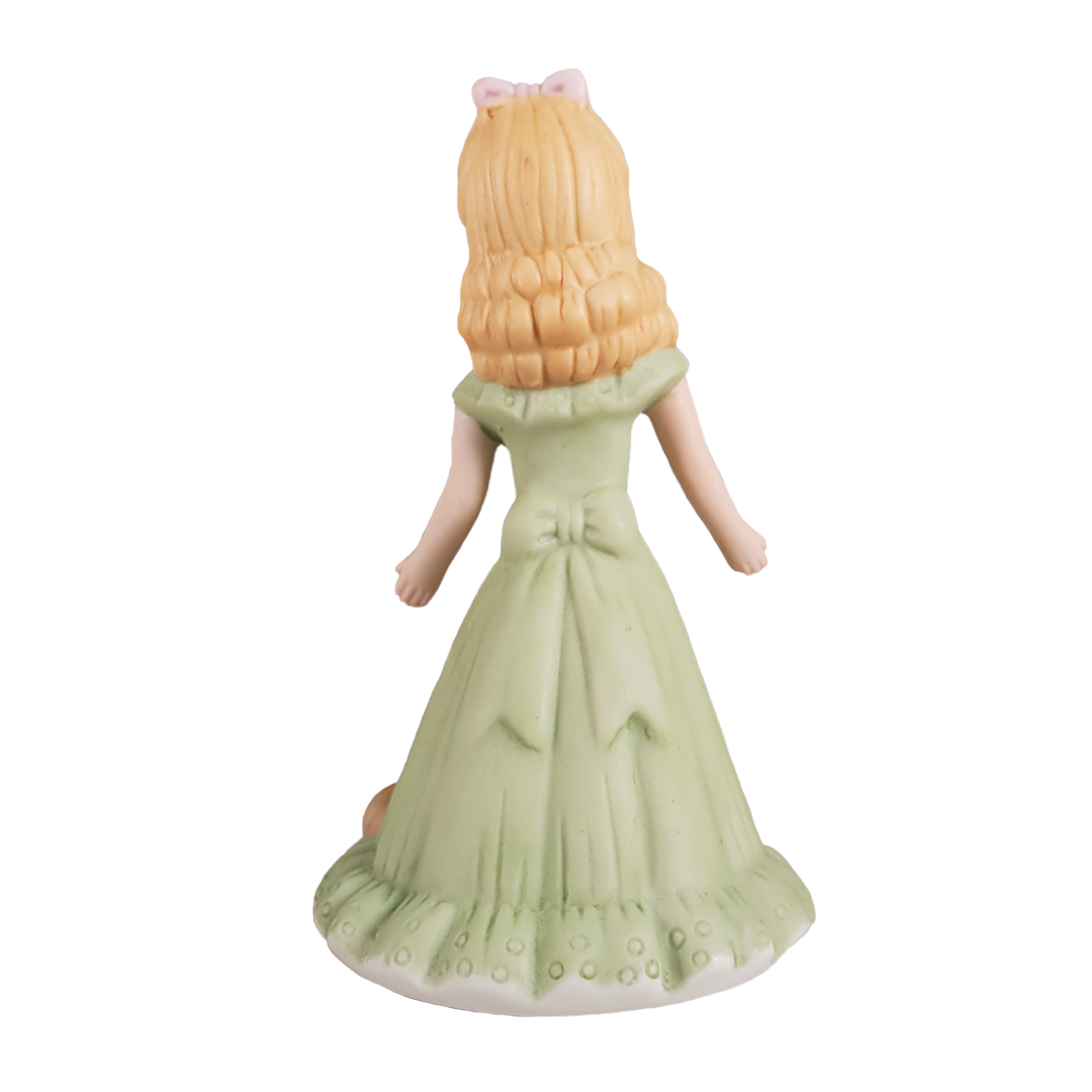 Back of figurine of a doll with blonde hair in a green dress on a white background