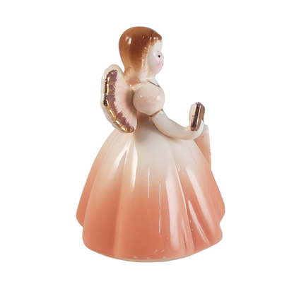 Side of porcelain angel figurine with a peach gradient dress holding a book on a white background